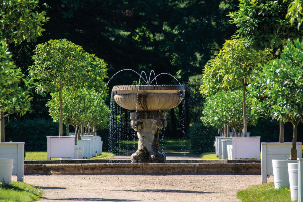 Fontaine en pierre à plusieurs vasques sur une allée engazonnée bordée d’arbres et de bacs blancs