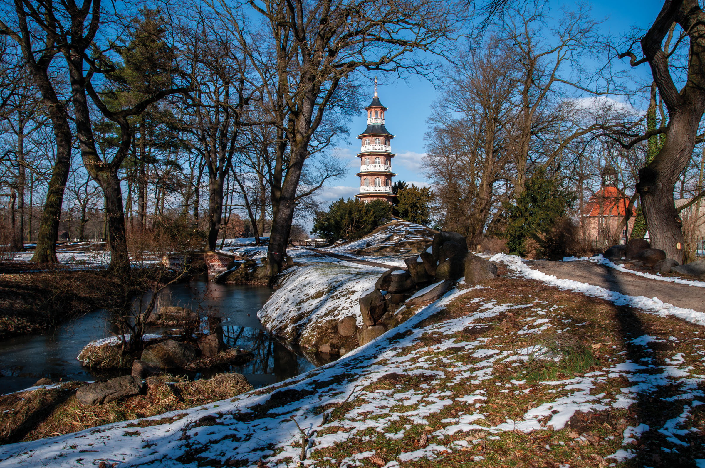 Pagoda bianca con più tetti ricurvi tra alberi spogli, neve al suolo e un corso d’acqua con rocce in primo piano