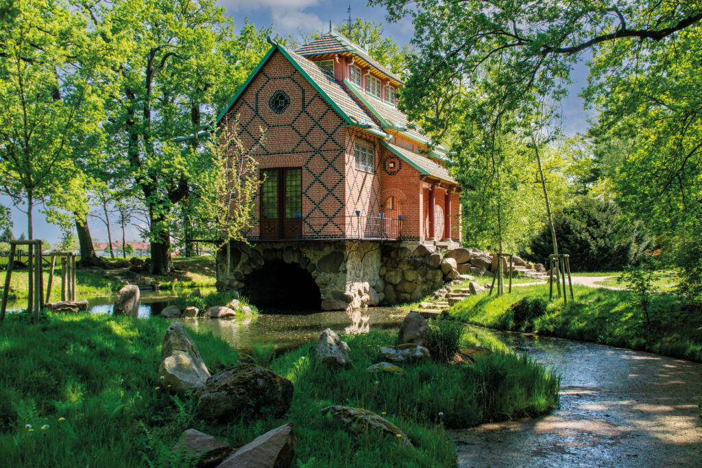 Maison de jardin à colombages rouges avec une petite tourelle sur un socle rocheux au-dessus d’un fossé, entourée d’arbres